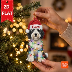 May include: A flat 2D Christmas tree ornament shaped like a black and white dog wearing a red Santa hat with the name Coco printed in white script. The dog is wrapped in a string of colorful Christmas lights with small glowing bulbs.
