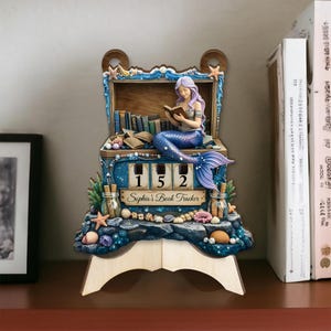 May include: A decorative mermaid book tracker with a blue and purple color scheme. The mermaid is reading a book, and the tracker displays the number 152. The text "Sophia's Book Tracker" is also visible.