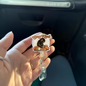 May include: A white and clear retractable badge reel featuring a cartoon graphic of a black creature on an orange pumpkin. The badge reel has the words "Stay Kind" in a brown script font.