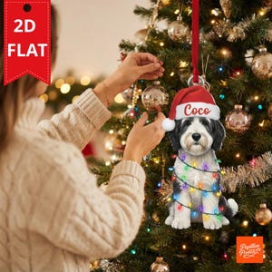 Op de afbeelding: Een platte, tweedimensionale kerstboomversiering met een zwart-witte hond die een rode kerstmuts draagt met de naam Coco in witte letters. De hond is omwikkeld met een snoer van kleurrijke, oplichtende kerstverlichting.