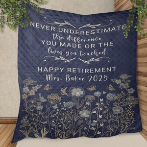 Puede incluir: Manta acolchada azul marino con texto blanco: "NUNCA SUBESTIMES la diferencia QUE HICISTE O las vidas que tocaste. FELIZ JUBILACIÓN Sra. Baker 2025." Presenta diseños florales y mariposas en dorado y azul claro.
