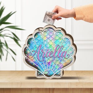 Puede incluir: Una hucha de madera con forma de concha marina con un diseño de escamas de sirena coloridas. El nombre "Ariella" está escrito en rosa. Una mano está insertando un billete de cien dólares. La hucha está sobre una superficie marrón claro.