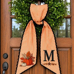 May include: A fabric door hanger featuring orange and white vertical stripes with a black scalloped edge. One side displays an embroidered pumpkin and autumn leaves, while the other side features a large black letter M above a small star and leaf motif.