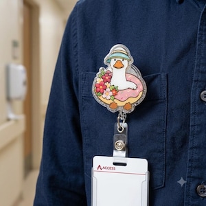 May include: A badge reel featuring a white duck wearing a light blue sun hat, floating in a pink donut decorated with pink and red flowers. The acrylic piece has a glittery finish and is attached to a clear plastic strap holding a white ID card.