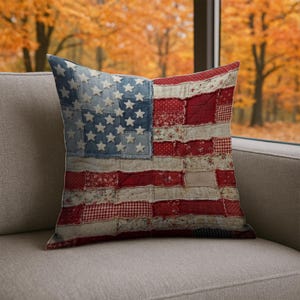 May include: A square decorative pillow with a patchwork design resembling the American flag. The pillow features a blue denim section with white stars and red and white striped sections. The pillow is placed on a gray sofa.