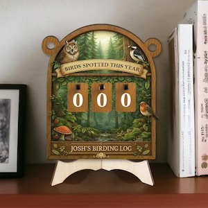 May include: Wooden birding log with a forest scene background. The log features a counter with the numbers "000" and the text "Birds Spotted This Year" and "Josh's Birding Log". Decorative birds and a mushroom are also featured.