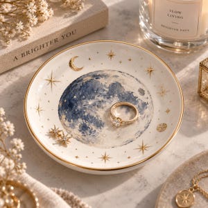 Puede incluir: Un plato decorativo redondo de cerámica blanca con una ilustración detallada de una luna azul estilo acuarela en el centro, acentuado con estrellas y una luna creciente en tono dorado. Un anillo dorado con una piedra clara descansa dentro.