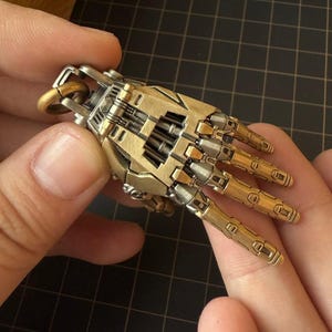 Peut inclure: Une miniature métallique détaillée d'une main robotique, avec des détails mécaniques complexes. La main est principalement de couleur or et argent, avec des doigts articulés et un joint de poignet complexe. Le design suggère un thème steampunk ou science-fiction.
