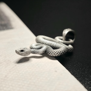 May include: A silver snake pendant with intricate scale detailing. The snake is coiled, with its head raised, and has a loop for a chain. The pendant is set against a contrasting black and white background. Likely made of metal.