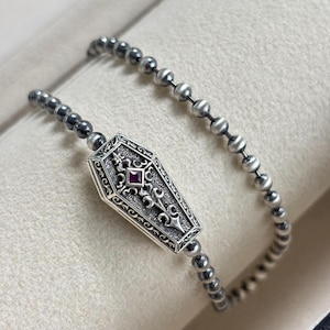 May include: A silver bracelet featuring a coffin-shaped charm with ornate detailing and a central red gemstone. The bracelet is composed of small, round beads in varying shades of gray and silver, creating a gothic or alternative aesthetic.