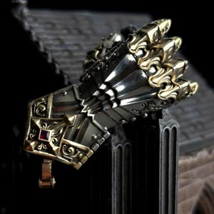 Puede incluir: Un guantelete detallado, de color plateado, con detalles dorados. El guantelete presenta diseños ornamentados, incluyendo una joya roja y patrones intrincados. Los dedos están rematados con formas doradas en forma de garra.