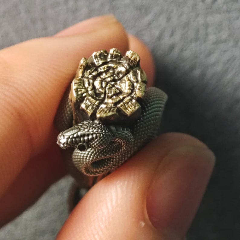 May include: A silver-coloured ring with a coiled snake design and textured body. The snake's head is raised, and a gold-coloured, ornate crown sits on its back. The ring is held between fingers against a neutral background.