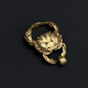 Può includere: Un ciondolo a forma di granchio color oro. Il granchio ha chele dettagliate e un guscio strutturato. Il ciondolo include un anello per il fissaggio a una catena o portachiavi. Lo sfondo è di colore scuro e uniforme.