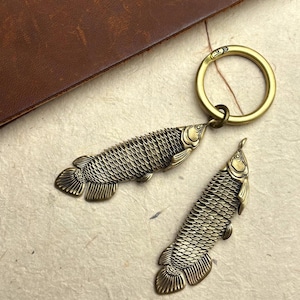 以下が含まれることがあります： 詳細な魚のペンダントが付いたブロンズ色のキーホルダー。魚は、円形のキーリングに取り付けられた、テクスチャ加工された鱗とひれを持っています。2つ目の魚のペンダントも表示されています。