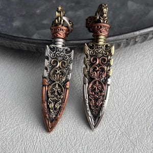 May include: Two ornate metal pendants with a dagger-like design. The pendants feature intricate patterns and a mix of silver, copper, and gold tones. Each pendant has a detailed lion figure at the top.