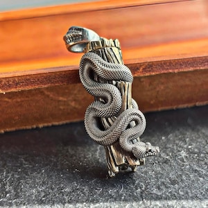 以下が含まれることがあります： 木製の柱に巻き付いたヘビを描いた、詳細なシルバーペンダント。ペンダントの上部には頭蓋骨があります。ヘビの鱗は細かく詳細です。暗い背景。ユニークなジュエリーです。