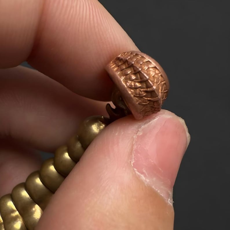 May include: A metallic object featuring a cylindrical top piece with a textured, scale-like pattern in a copper tone, connected to a series of smooth, rounded, gold-toned spherical segments that form a chain-like structure.