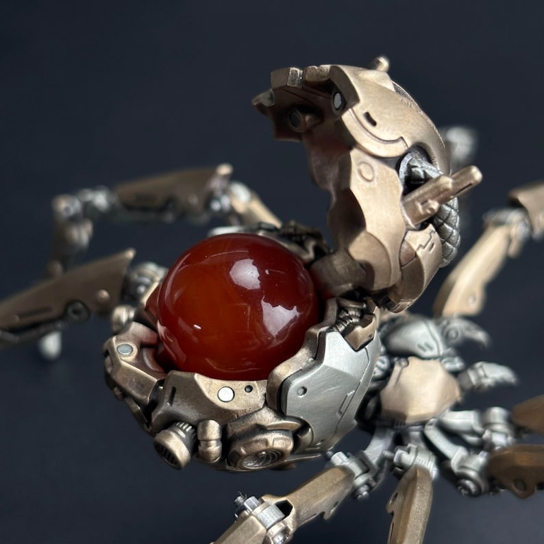 May include: Detailed close-up of a mechanical spider-like sculpture. The sculpture features a metallic gold and silver color scheme with intricate details. A large, polished, reddish-orange sphere is held within the sculpture's open compartment, creating a striking contrast.