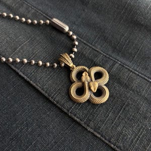 Op de afbeelding: Een ketting met een messing hanger met een gestileerd slangenmotief. De hanger heeft de vorm van een klavertjevier, waarbij elk blad door een slang wordt gevormd. De ketting heeft een zilverkleurige kogelketting.