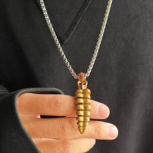 May include: A metallic gold-toned pendant shaped like a rattlesnake rattle, suspended from a silver-colored box chain necklace. The pendant features a segmented, tapered design with a textured surface, hanging vertically from a small metal bail.