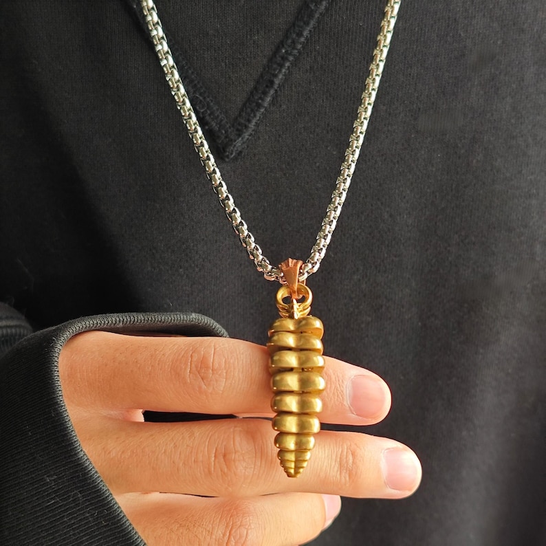 May include: A metallic gold-toned pendant shaped like a rattlesnake rattle, suspended from a silver-colored box chain necklace. The pendant features a segmented, tapered design with a textured surface, hanging vertically from a small metal bail.