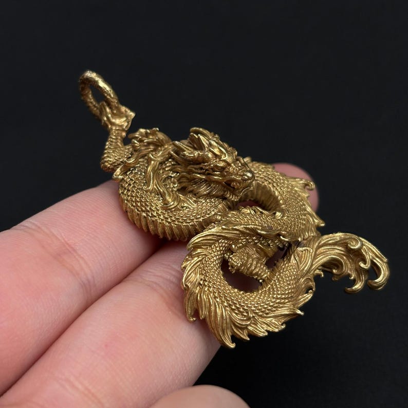 May include: A detailed, golden dragon pendant. The dragon is intricately sculpted with scales, a flowing mane, and curled tail. The pendant is suspended from a loop at the top, suggesting it's designed to be worn as jewelry. The background is a solid black.