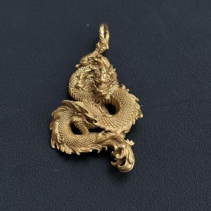 May include: A gold-colored dragon pendant with intricate detailing. The dragon is coiled, with detailed scales, a flowing mane, and a loop at the top for a chain. The pendant is set against a dark background.