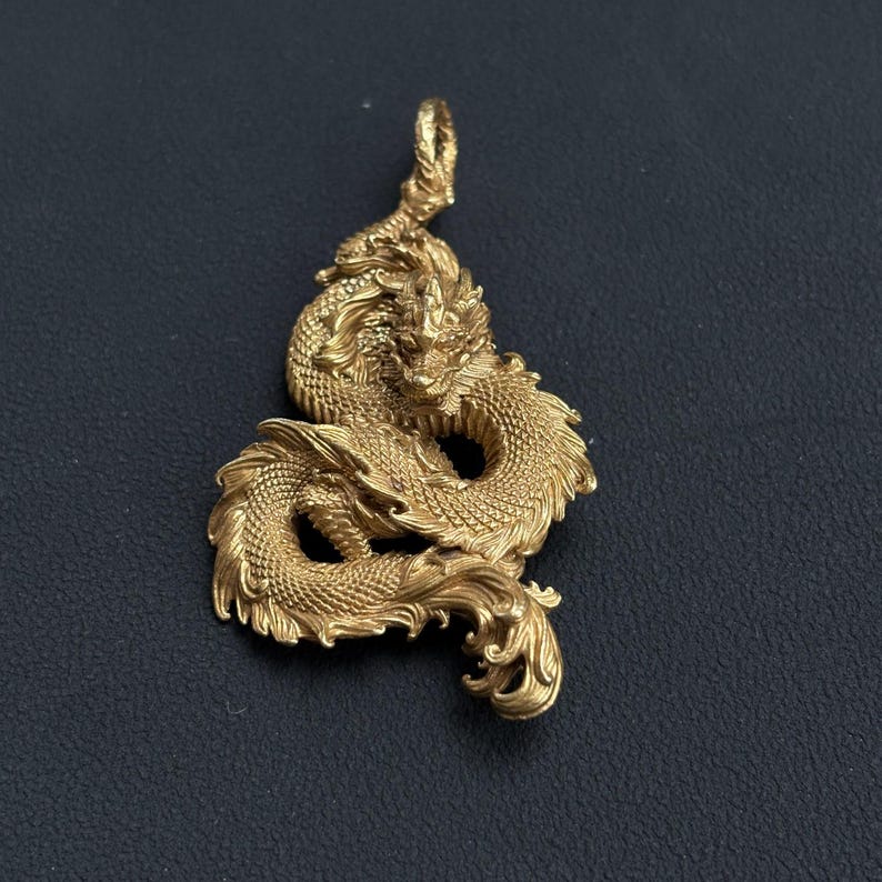 May include: A gold-colored dragon pendant with intricate detailing. The dragon is coiled, with detailed scales, a flowing mane, and a loop at the top for a chain. The pendant is set against a dark background.