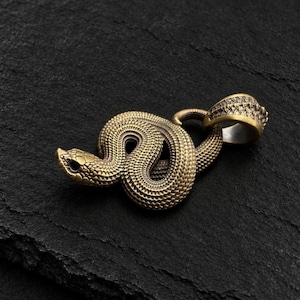 May include: A brass-toned metal pendant shaped like a coiled snake with detailed scale textures across its body. The snake is positioned in a circular loop with its head facing left, and it features a sturdy integrated bail at the top.