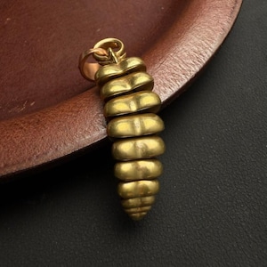 May include: A gold-colored pendant with a spiral design. The pendant is made of multiple stacked, heart-shaped segments, creating a textured, elongated shape. It has a small loop at the top for attaching to a chain. The pendant is displayed on a brown leather surface.