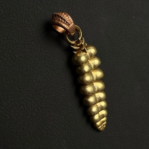 May include: A gold-toned pendant shaped like a rattlesnake rattle with a segmented, tapered body. The top features a textured, copper-colored bail loop for attachment. The surface has a smooth, metallic finish with distinct horizontal ridges.