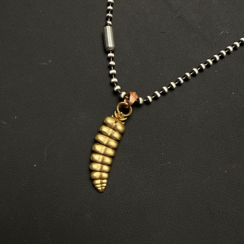 May include: A metallic ball chain necklace featuring a gold-toned pendant shaped like a segmented rattlesnake tail. The pendant is attached to the chain by a small circular ring and a copper-colored connector, creating a distinct textured appearance.
