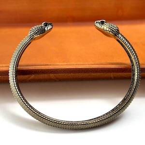 May include: A gold-toned metal cuff bracelet featuring a textured snake scale pattern throughout the band. The two ends of the open-style bracelet are shaped like detailed snake heads with dark, recessed eyes and defined facial features.