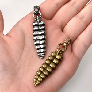 May include: Two rattlesnake rattle pendants, one in a polished silver-tone metal and one in a brass-tone metal. Each pendant features a segmented, tapered design with a textured bail at the top for attachment to a necklace chain or cord.