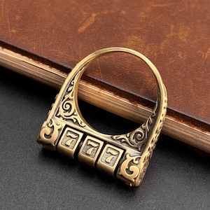 May include: A brass-toned metal ring featuring an integrated three-digit combination lock mechanism. The band is decorated with ornate scrollwork engravings and displays the number 777 on the rotating tumblers at the base of the ring.