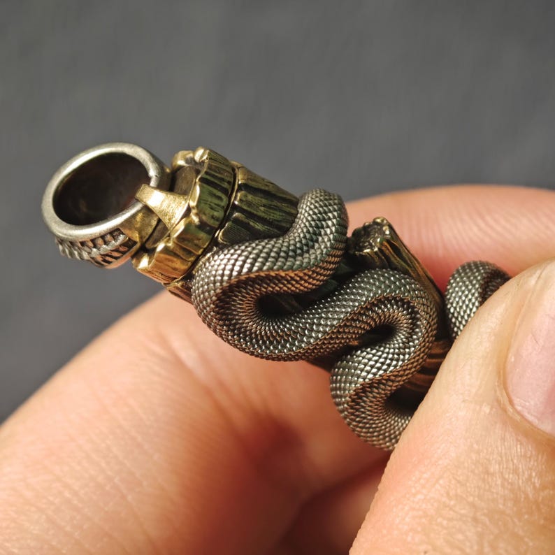 May include: A close-up of a metal bottle stopper. The stopper features a silver ring, a gold-coloured cylindrical section, and a textured snake wrapped around the body. The snake has a scale-like pattern.