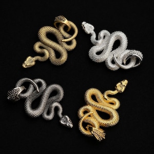 May include: Four snake-shaped pendants arranged in a square formation. Each pendant features a textured, scaled surface and a coiled body. The set includes one matte gold, one silver, one dark gunmetal, and one bright yellow gold snake pendant.