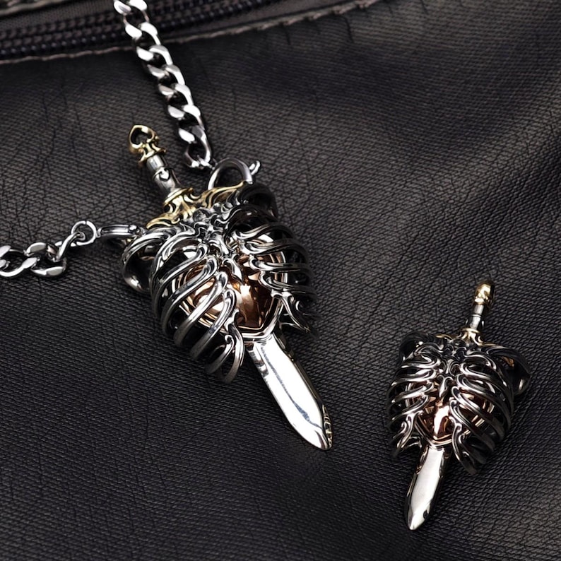 Op de afbeelding: Een zilverkleurige metalen ketting met hanger en een bijpassende losse hanger, beide voorzien van een gedetailleerde anatomische ribbenkaststructuur rondom een centraal hart, doorboord door een zwaard dat verticaal door het ontwerp loopt.