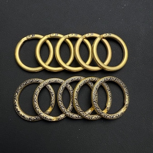 Könnte beinhalten: Eine Sammlung von acht goldfarbenen Metallringen. Die obere Reihe zeigt schlichte Ringe, während die untere Reihe Ringe mit einem schwarz-goldenen Dekorationsmuster aufweist. Die Ringe sind auf einer dunklen Oberfläche angeordnet.