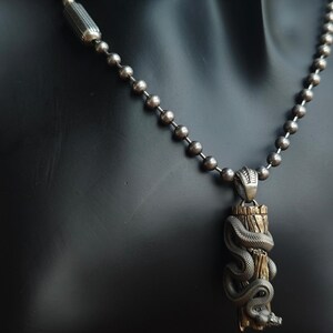 May include: A silver-coloured necklace with a pendant depicting a detailed snake coiled around a wooden post. The snake's head is visible at the bottom, and the necklace has a ball chain design.