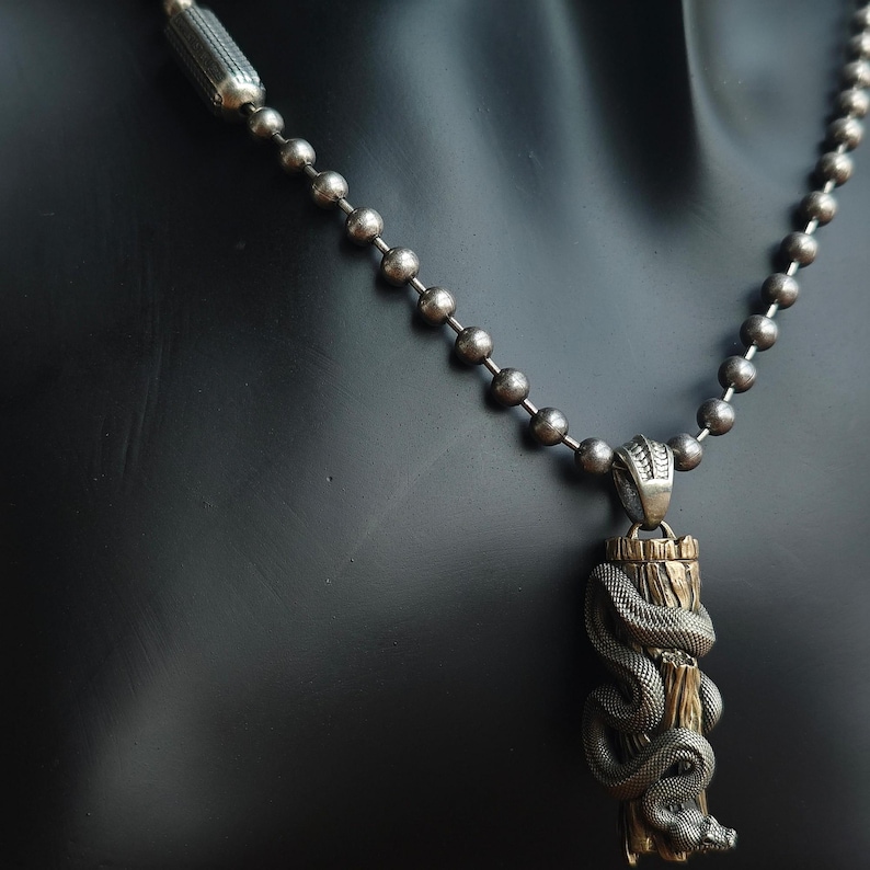 May include: A silver-coloured necklace with a pendant depicting a detailed snake coiled around a wooden post. The snake's head is visible at the bottom, and the necklace has a ball chain design.