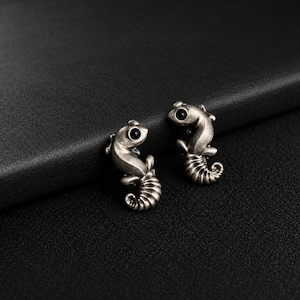 Puede incluir: Un par de pendientes de botón con forma de caballito de mar con acabado en plata envejecida y pequeñas piedras circulares negras como ojos. Las figuras de caballito de mar presentan colas curvas detalladas y cuerpos texturizados de metal.