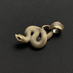 May include: A bronze-toned snake pendant with a textured body and a ring bail. The snake is coiled, with its head raised. The pendant has a vintage or antique aesthetic.