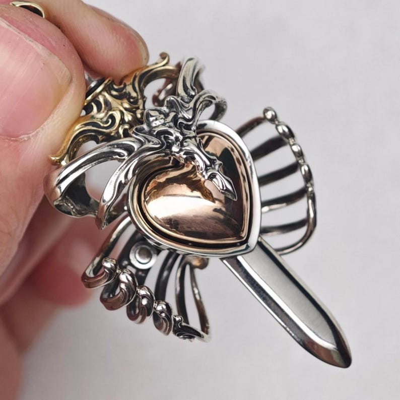 Op de afbeelding: Een metalen hanger met een centraal gepolijst hartvormig element in ros&eacute;goud, bedekt met ingewikkeld zilverkleurig filigraan. Het ontwerp bevat een puntig, zwaardachtig element dat naar beneden uitsteekt, geaccentueerd met goudkleurige details.