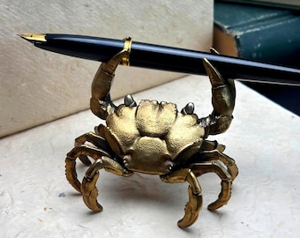 May include: A brass crab pen holder with a black pen. The crab is detailed with a golden finish and holds the pen with its claws. The pen has a gold-colored nib and accents.
