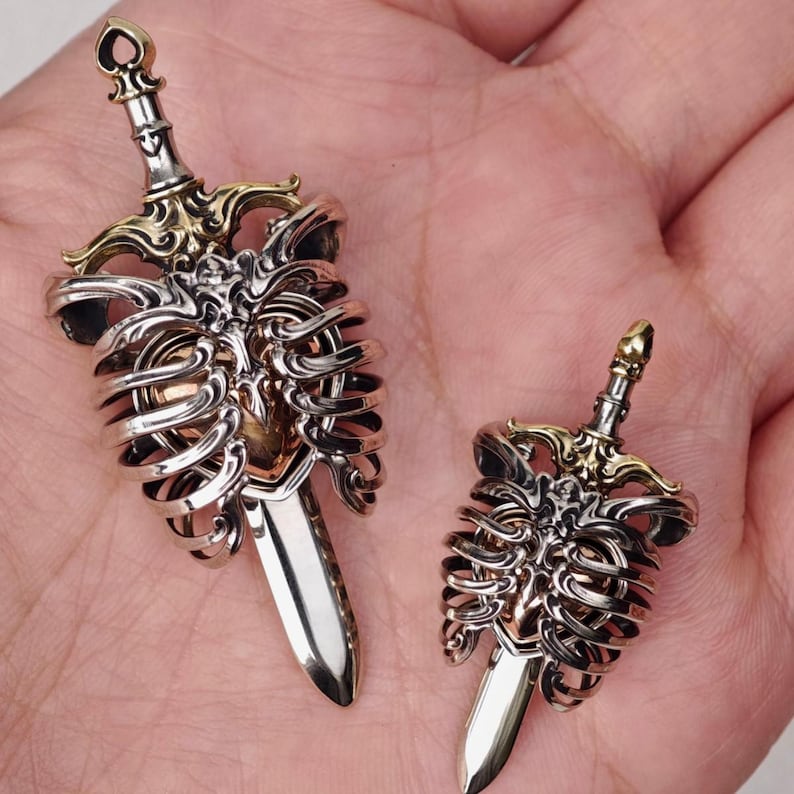 Op de afbeelding: Twee hangers van zilver- en goudkleurig metaal, elk in de vorm van een zwaard dat door een menselijke ribbenkast steekt. De ontwerpen bevatten gedetailleerde, gepolijste metalen elementen met een hartvormig middenstuk tussen de ribben.