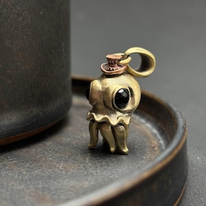 May include: A brass octopus pendant with a small top hat and black gemstone eyes. The octopus has a detailed design with tentacles and a loop for a necklace chain. The pendant is on a dark surface.