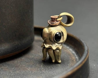 May include: A brass-toned metal pendant shaped like a small octopus with large, smooth black eyes. The octopus wears a tiny, copper-colored top hat on its head, and a circular loop is attached to the top for use as a necklace charm or keychain.