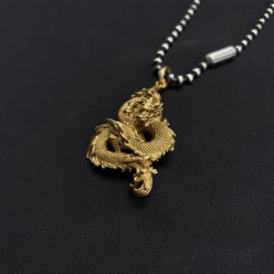 May include: A gold-colored dragon pendant necklace. The dragon is intricately detailed and suspended from a chain of alternating black and silver beads. The pendant is the focal point, showcasing the mythical creature's scales and form.
