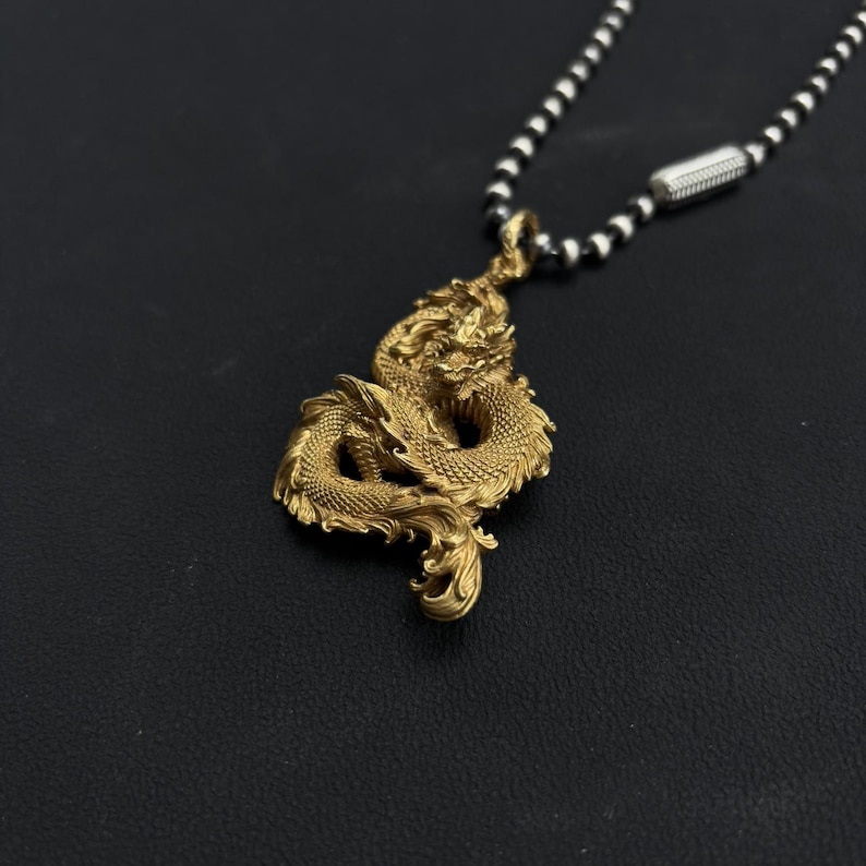 May include: A gold-colored dragon pendant necklace. The dragon is intricately detailed and suspended from a chain of alternating black and silver beads. The pendant is the focal point, showcasing the mythical creature's scales and form.